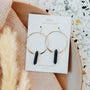 A pair of Smoky Quartz Hoop - Protection earrings with handcrafted gold hoops and black smoky quartz pendants, displayed on a branded card atop a neutral plate with blush fabric and dried pampas grass in the background.