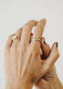 Two light-skinned hands are gently clasped on a white background. The left wears an 18k gold-plated flower ring; the right features the White Opal Inset Baguette ring. Nails are short, dark-painted.