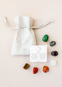 A white drawstring pouch labeled “Chakra Crystal Kit” sits next to an info card about chakra healing crystals, with eight colorful polished stones from the kit arranged around the pouch and card on a light surface.