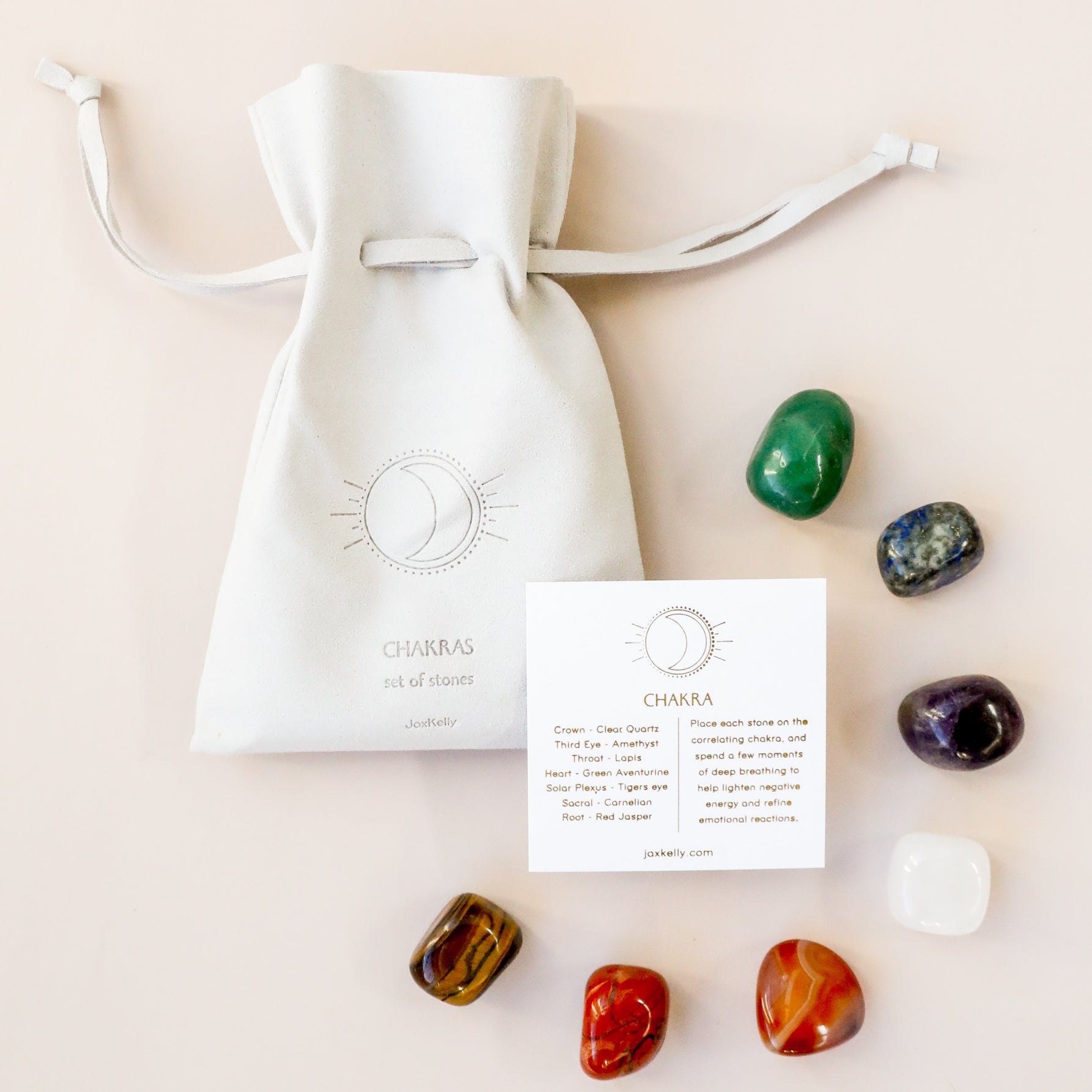 A white drawstring pouch labeled “Chakra Crystal Kit” sits next to an info card about chakra healing crystals, with eight colorful polished stones from the kit arranged around the pouch and card on a light surface.