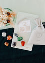 The Chakra Crystal Kit includes six colorful chakra healing crystals, a white CHAKRAS pouch, an info card, and dried flowers, all arranged on a notebook and dark cloth.