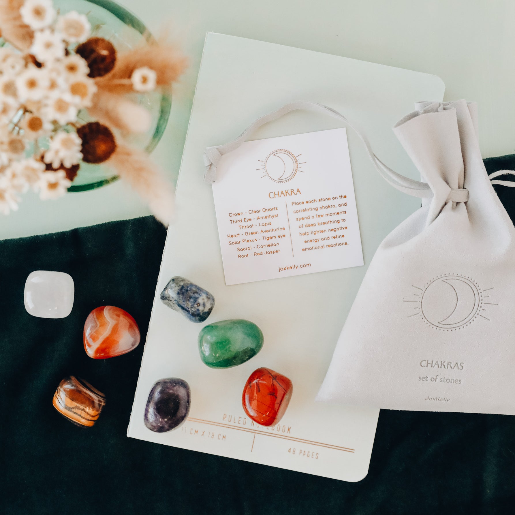 The Chakra Crystal Kit includes six colorful chakra healing crystals, a white CHAKRAS pouch, an info card, and dried flowers, all arranged on a notebook and dark cloth.