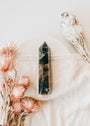 A Labradorite Crystal Point rests on a light cloth, surrounded by dried pink flowers and a bundle of white sage, creating a serene, protective display against a soft, neutral background.