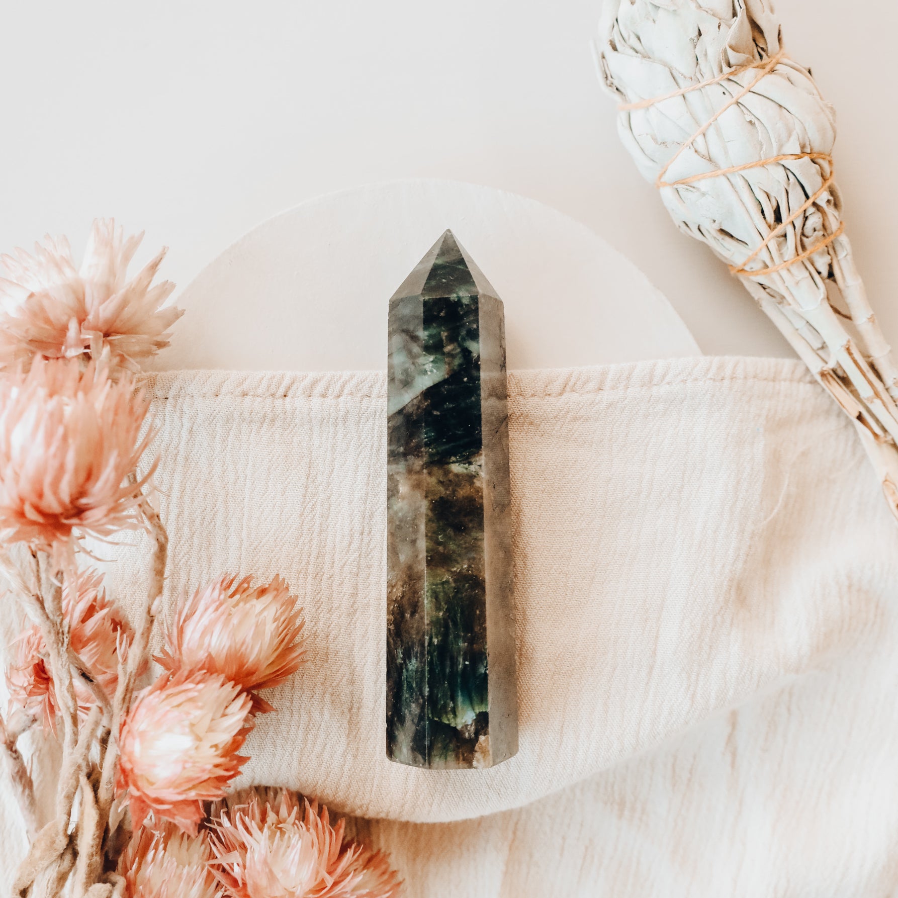 A Labradorite Crystal Point rests on a light cloth, surrounded by dried pink flowers and a bundle of white sage, creating a serene, protective display against a soft, neutral background.