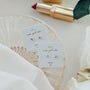 A glass dish holds two earring cards, one with pink studs and the other featuring “Tiny White Opal Heart” lab-grown white opal studs. A gold lipstick and white flowers rest nearby on a light surface.