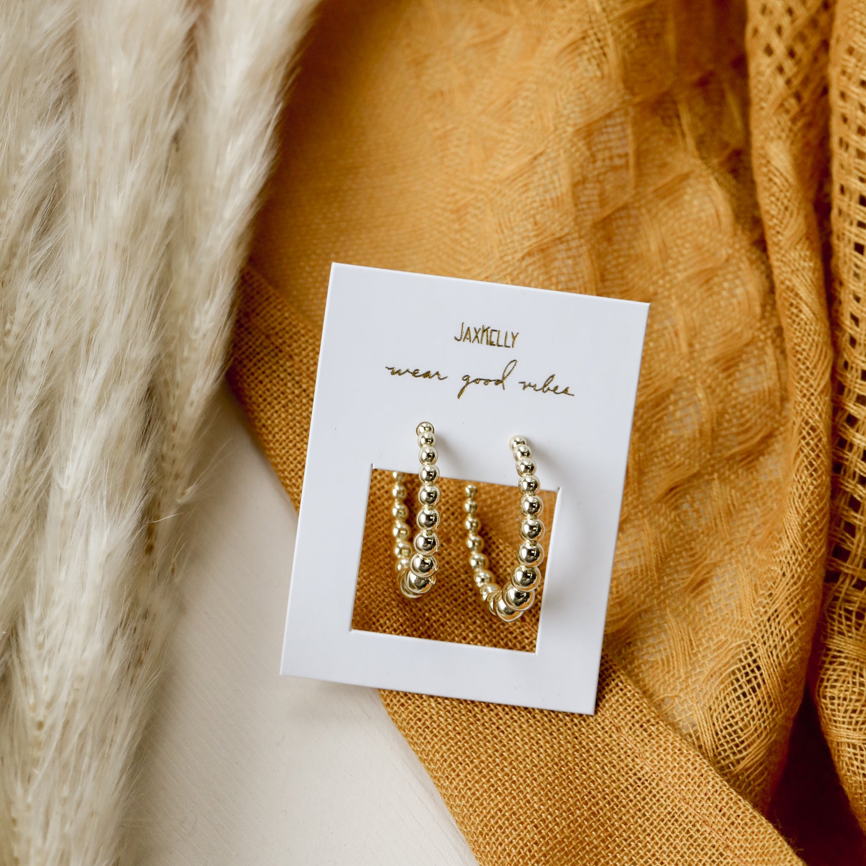 The Large Ball Hoop features gold-plated hoops with sterling silver posts, displayed on a white JaxKelly card that reads "wear good vibes," set atop a soft surface with beige pampas grass and mustard yellow textured fabric.