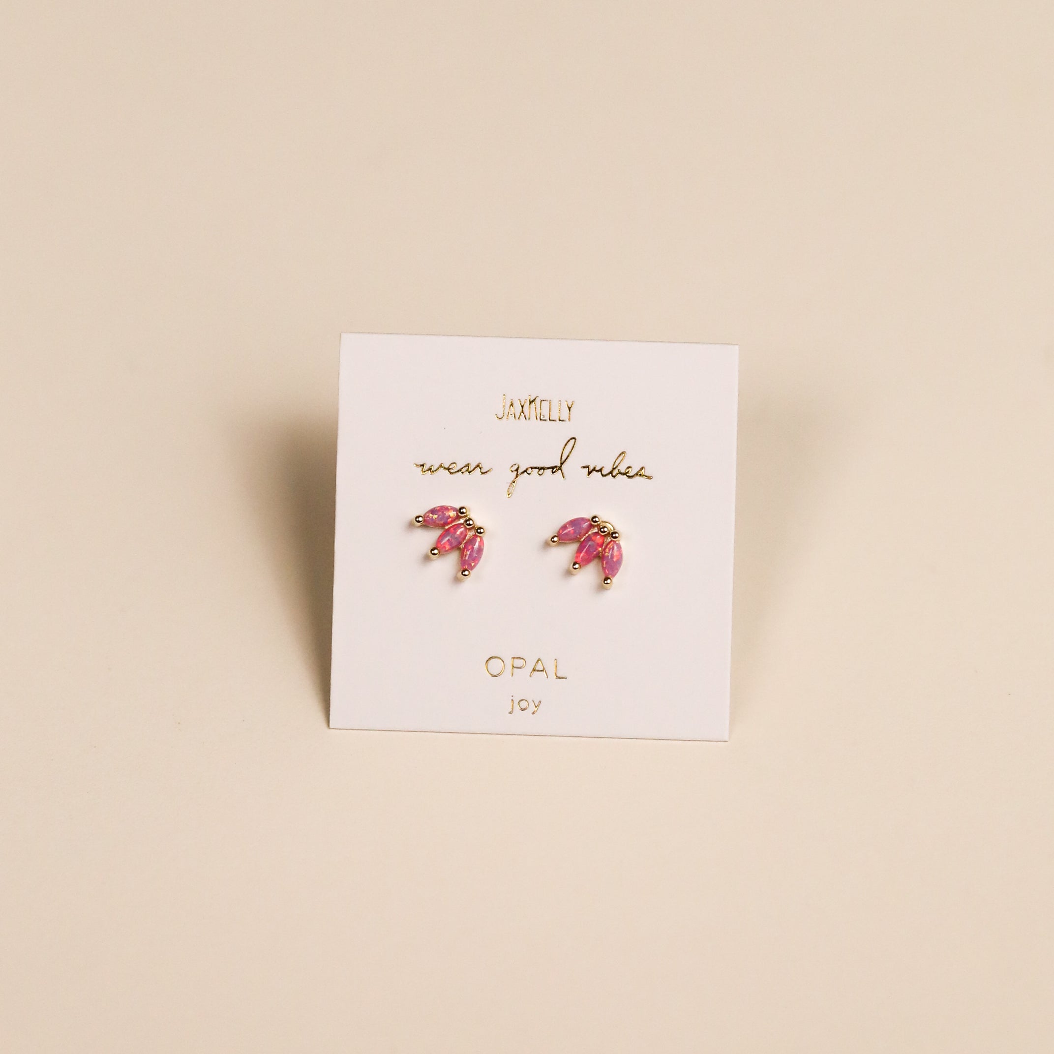 The Pink Opal Crown earrings, shaped like pink crescent moons with lab-grown Pink Opal and 18k gold plating, are shown on a white JAXKELLY wear good vibes OPAL joy card, set against a beige background.