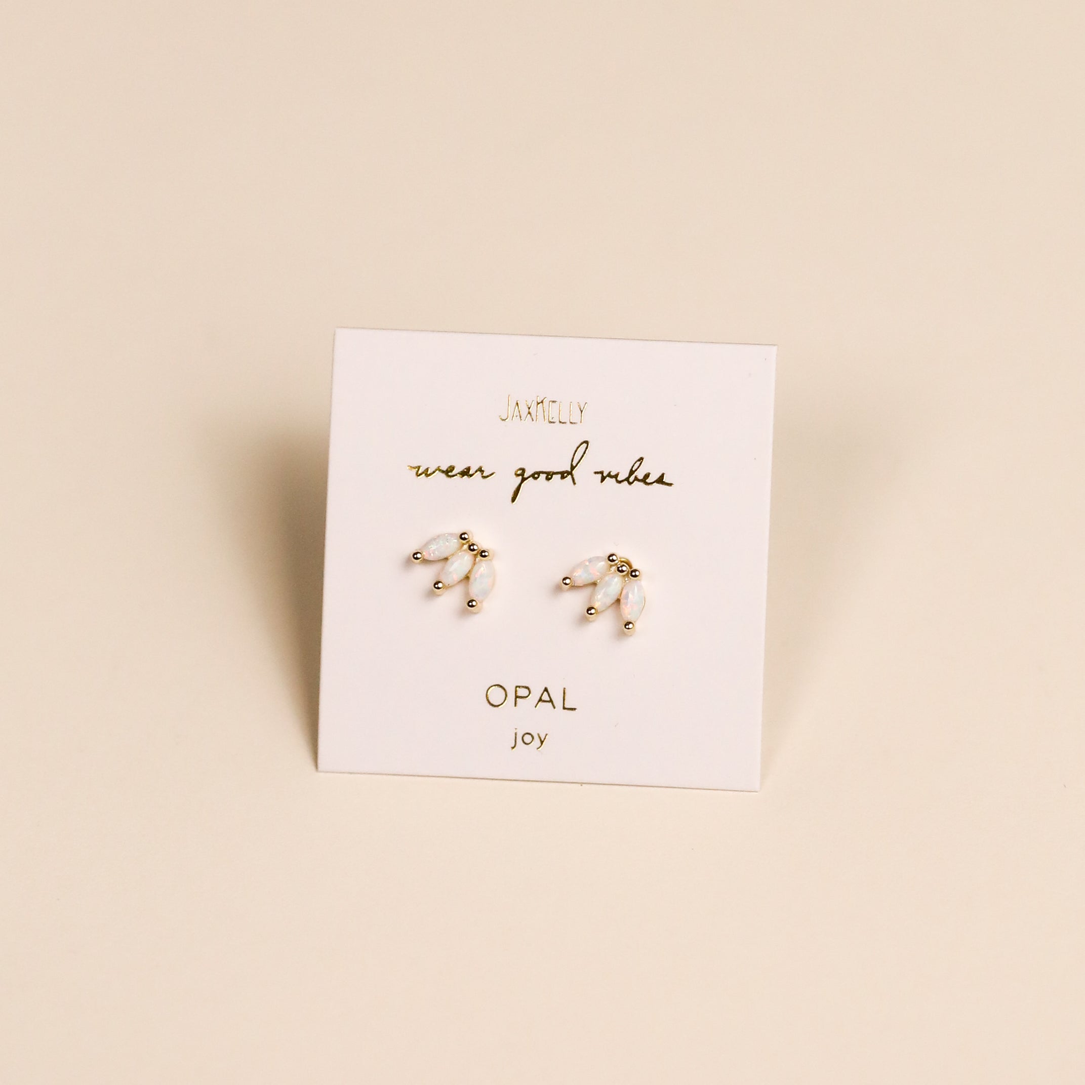 A pair of delicate gold and pearl White Opal Crown Stud earrings rest on a white JaxKelly card reading "wear good vibes" and "lab-grown opal joy," set against a neutral beige background.