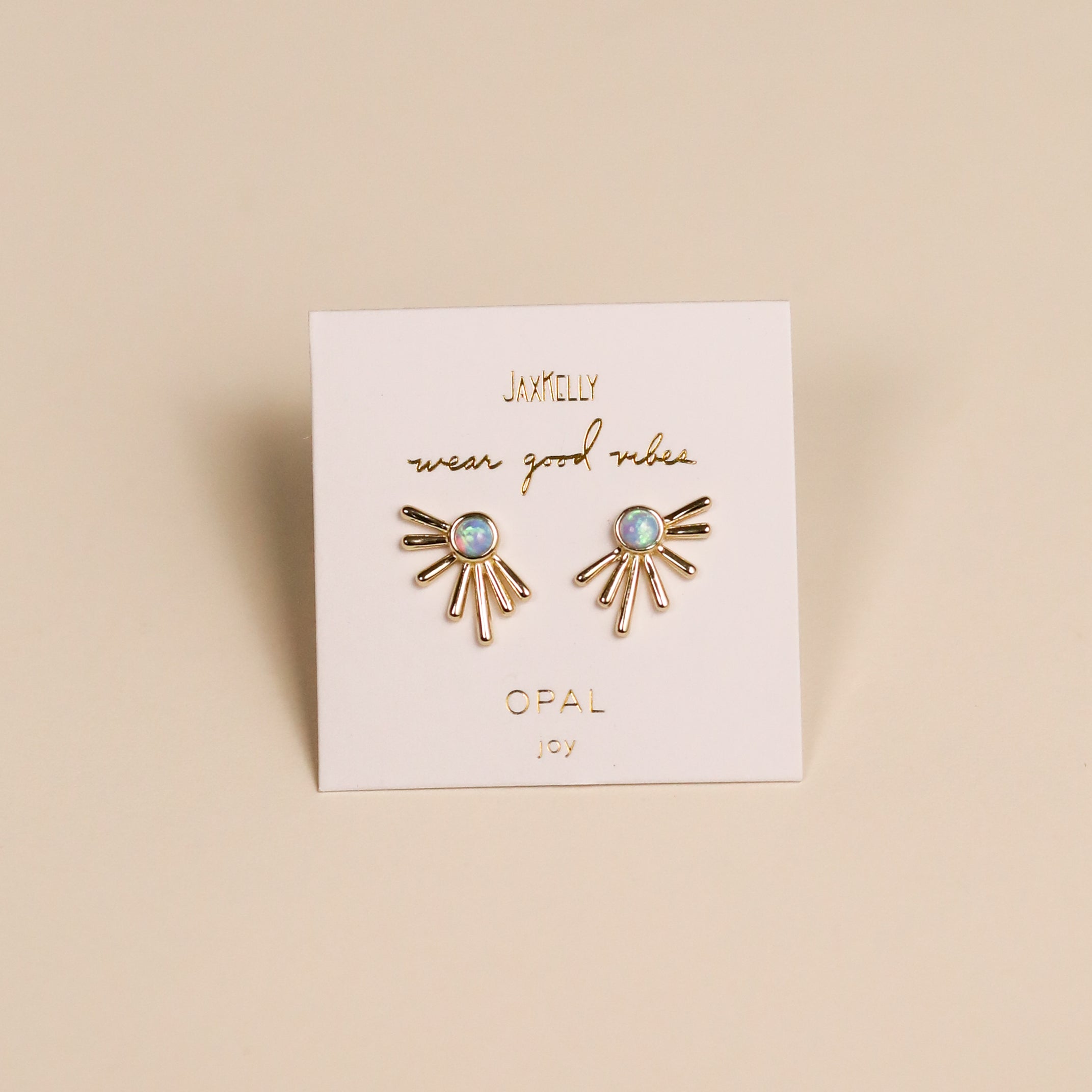 A pair of Sun Ray - Fire Opal stud earrings with lab-grown Fire Opal and 18k gold plating, shaped like sunbursts, displayed on a white JAXKELLY card reading "wear good vibes, OPAL, joy" against a beige background.