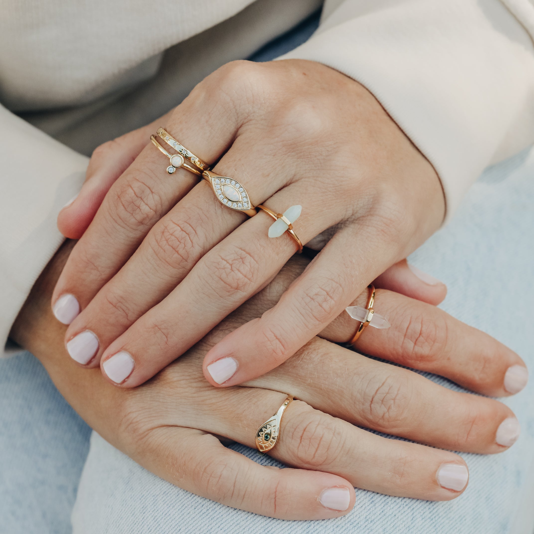 Close-up of hands with light pink nails wearing the Amazonite Point Ring and other gold rings with opal and white stones—an ideal minimalist jewelry look—resting on light blue jeans and a cream long sleeve top.