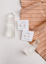 A pair of Rose Quartz Mineral Point - Love earrings with polished gemstones sit on a white card atop peach textured fabric, accented by large clear quartz crystals. Each earring features sterling silver with 18k gold plating for a refined finish.