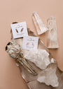 A flat lay image showcases the Rose Quartz Mineral Point - Love, paired with clear quartz crystals, gemstone earrings, and a bundle of sage on a metallic leaf atop a beige background.