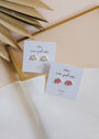 Two pairs of Pink Opal Crown stud earrings with lab-grown white stones and pink opal, set in 18k gold plating, are displayed on cards against a white cloth background with dried palm leaves in the corner.