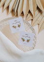 Two pairs of Sun Ray - Fire Opal gold earrings are displayed on white JaxKelly cards in a glass dish, surrounded by dried palm leaves and white fabric.