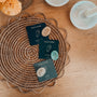 Three cards labeled Touch Stone with polished gemstones—Moonstone, Amazonite, and Chert—rest on a woven placemat atop a wooden table, accompanied by a flower, white decorative dishes, and the Citrine Touch Stone.