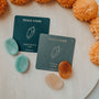 The Citrine Touch Stone features a polished natural citrine for success and a green Amazonite for hope, both displayed on labeled cards atop a light surface with bright orange flowers surrounding them.