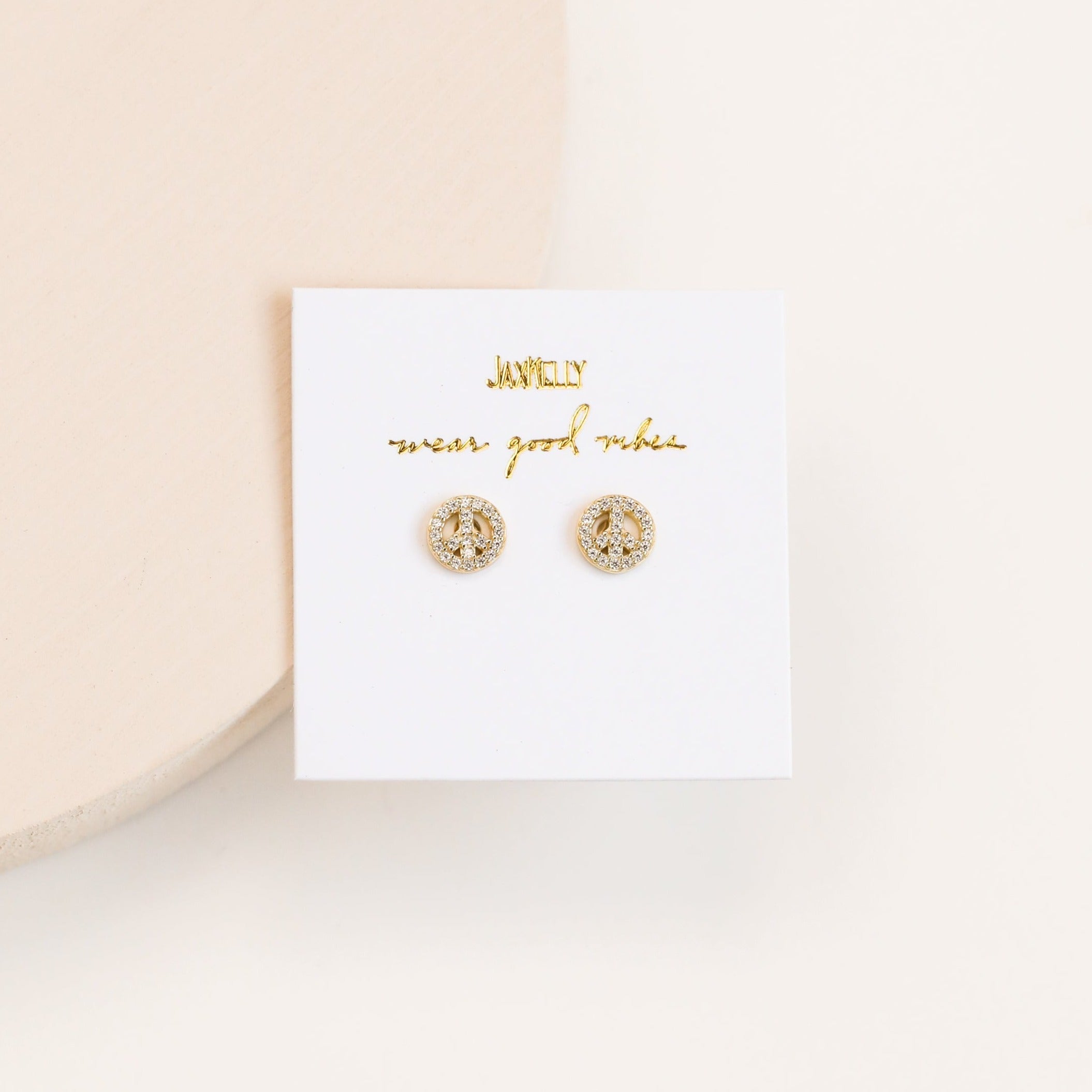 The Pavé Peace Sign earrings are small, round, 18k gold plated with embedded CZ stones, shown on a white card labeled "wear good vibes" in gold lettering.