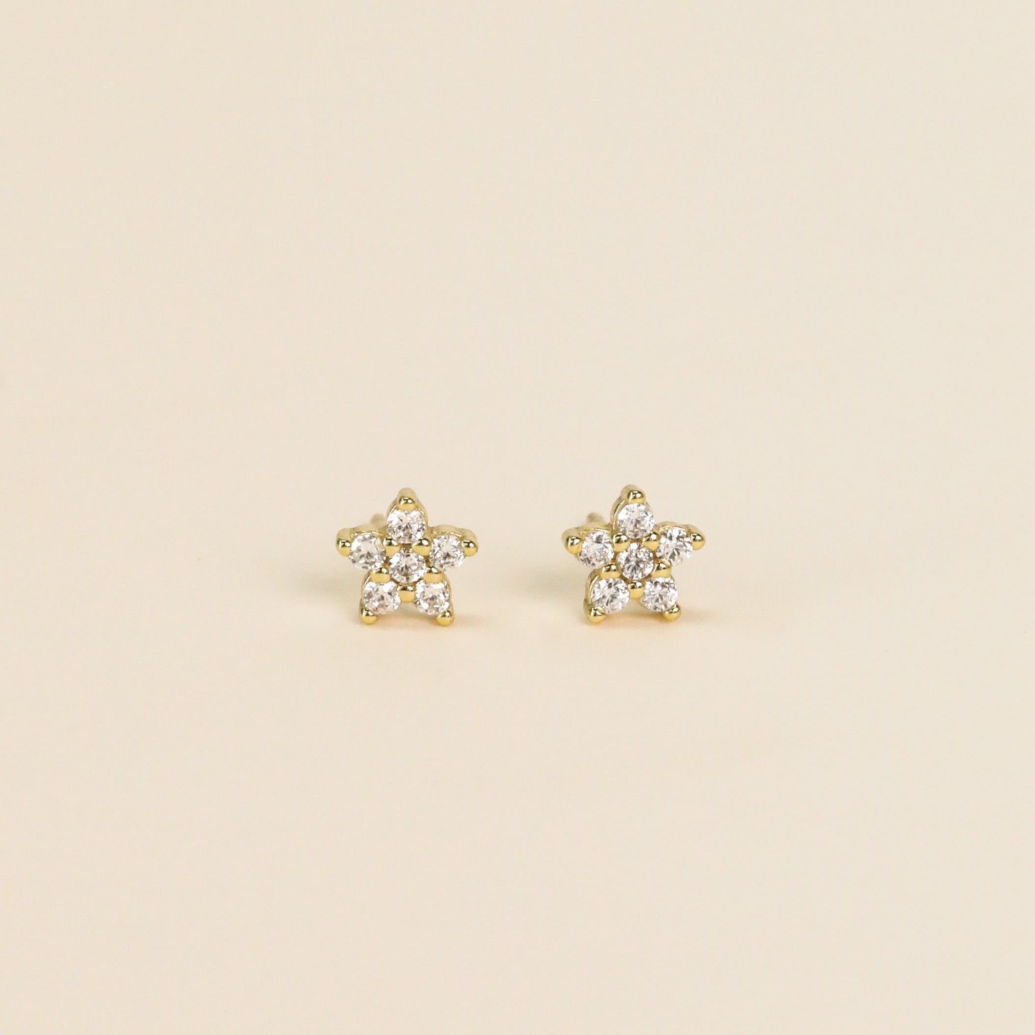 The Pavé Flower earrings feature star-shaped studs with 18k gold plated settings and sterling silver posts, each accented with sparkling clear gemstones. Shown on a simple, light beige background.