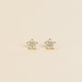 The Pavé Flower earrings feature star-shaped studs with 18k gold plated settings and sterling silver posts, each accented with sparkling clear gemstones. Shown on a simple, light beige background.