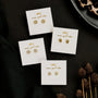 Four pairs of Pavé Flower gold stud earrings with 18k gold plating and sterling silver posts are displayed on white cards reading "never good rode" on a black background, accented by pinecones and part of a leopard-print dish.