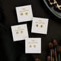 Four pairs of Pavé Peace Sign 18k gold plated earrings are displayed on white cards with gold writing, arranged on a dark surface with dried pinecones and animal print fabric in the background.