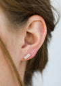 Close-up of a person’s ear with brown hair tucked behind it, wearing the White Bar Druzy—an 18k gold plated, small rectangular earring featuring two clear stones. The softly blurred background is neutral in color.