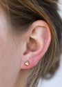 Close-up of a person's ear with light brown hair, wearing the Rose Quartz Mini Energy Gems - Love earring, a small triangular stud featuring rose quartz, 18k gold plating, and sterling silver posts.