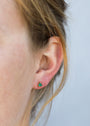 Close-up of a person’s ear with light brown hair, wearing Green Onyx Mini Energy Gems - Abundance: small green onyx triangular studs set in 18k gold plating. The background is plain and pale.