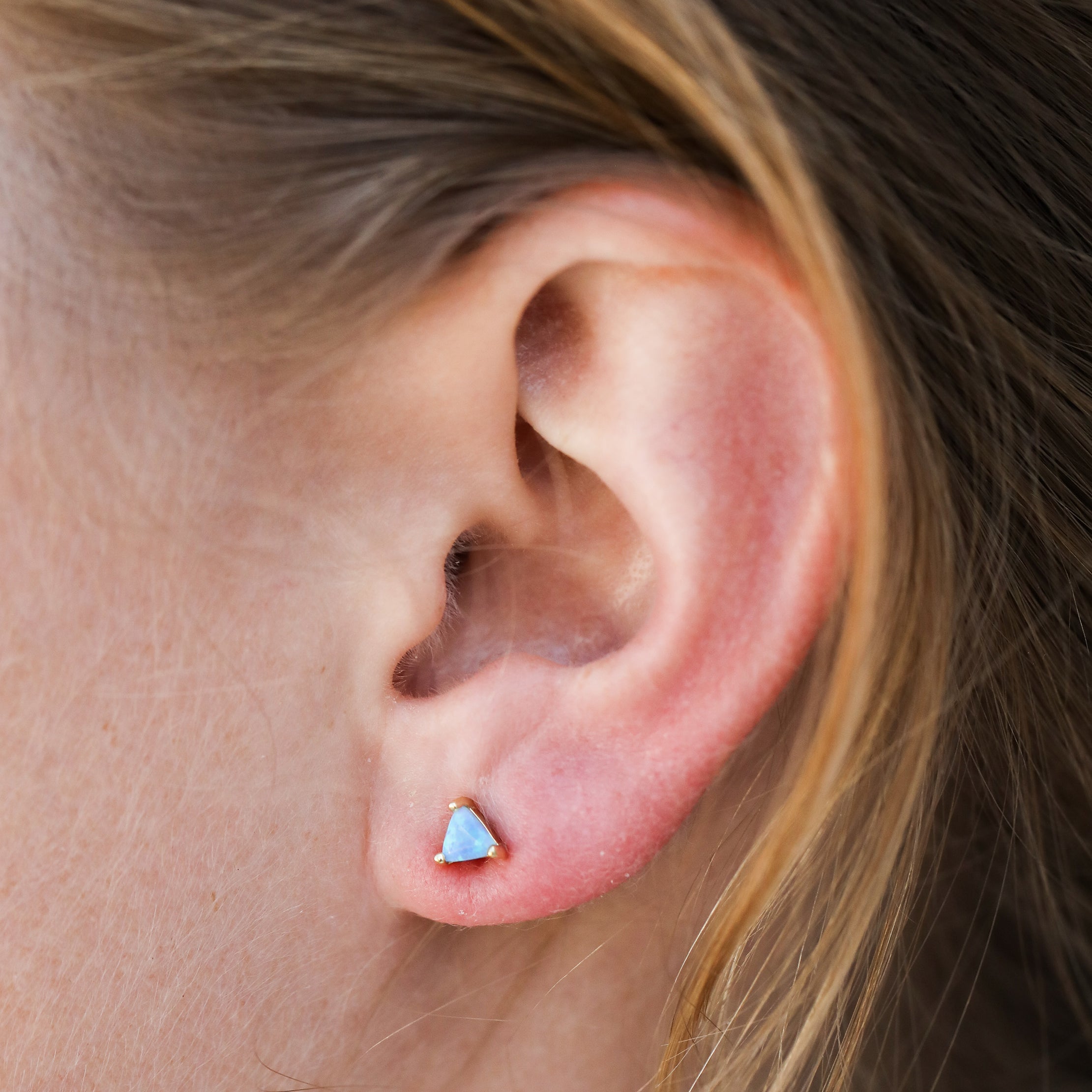 Close-up of a light-skinned person with light brown hair wearing the Fire Opal Mini Energy Gems - Joy stud, showcasing its small triangular design with lab-grown fire opal in elegant 18k gold plating.