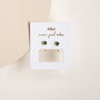 The Emerald CZ Huggie earrings feature emerald CZ stones and 18k gold plating, displayed on a white JaxKelly card reading "wears good vibes," set against a light, neutral background.