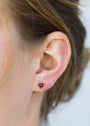 Close-up of a woman's ear wearing the Black Tourmaline Mini Energy Gems - Strength: small black triangular stud earrings with 18k gold plating and sterling silver posts. Her hair is tied back, background softly blurred.