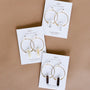 A pair of handcrafted crystal earrings—Smoky Quartz Hoop - Protection—is displayed on a white card against a brown background. The card highlights the stone name and features an uplifting message about protection.
