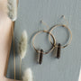 The Smoky Quartz Hoop - Protection earrings, featuring gold hoops with dark smoky quartz pendants, are displayed on a textured light blue surface beside soft white dried grass—an elegant showcase of handcrafted style.