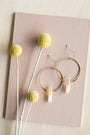 The JaxKelly Citrine Hoop - Success earrings, featuring gold hoops with clear crystal pendants, are elegantly displayed on a pink textured surface beside yellow billy button flowers, showcasing these hypoallergenic jewelry pieces.