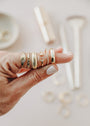 A hand with light nail polish showcases five unique gold rings—all stacked on the index finger—including the Celestial Ring. The soft beige background features blurred jewelry tools and extra rings.