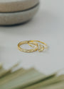 The Celestial Ring set features a gold band engraved with small stars and a crescent moon ring adorned with sparkling stones, resembling cubic zirconia. Both rest on a white surface with a blurred green leaf and ceramic dish in the background.