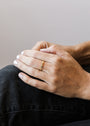 A close-up of a person’s hands on their knee, showing neatly manicured nails with white polish and the Celestial Ring—a simple gold stacking ring with delicate details—on one finger. The person is wearing dark pants.