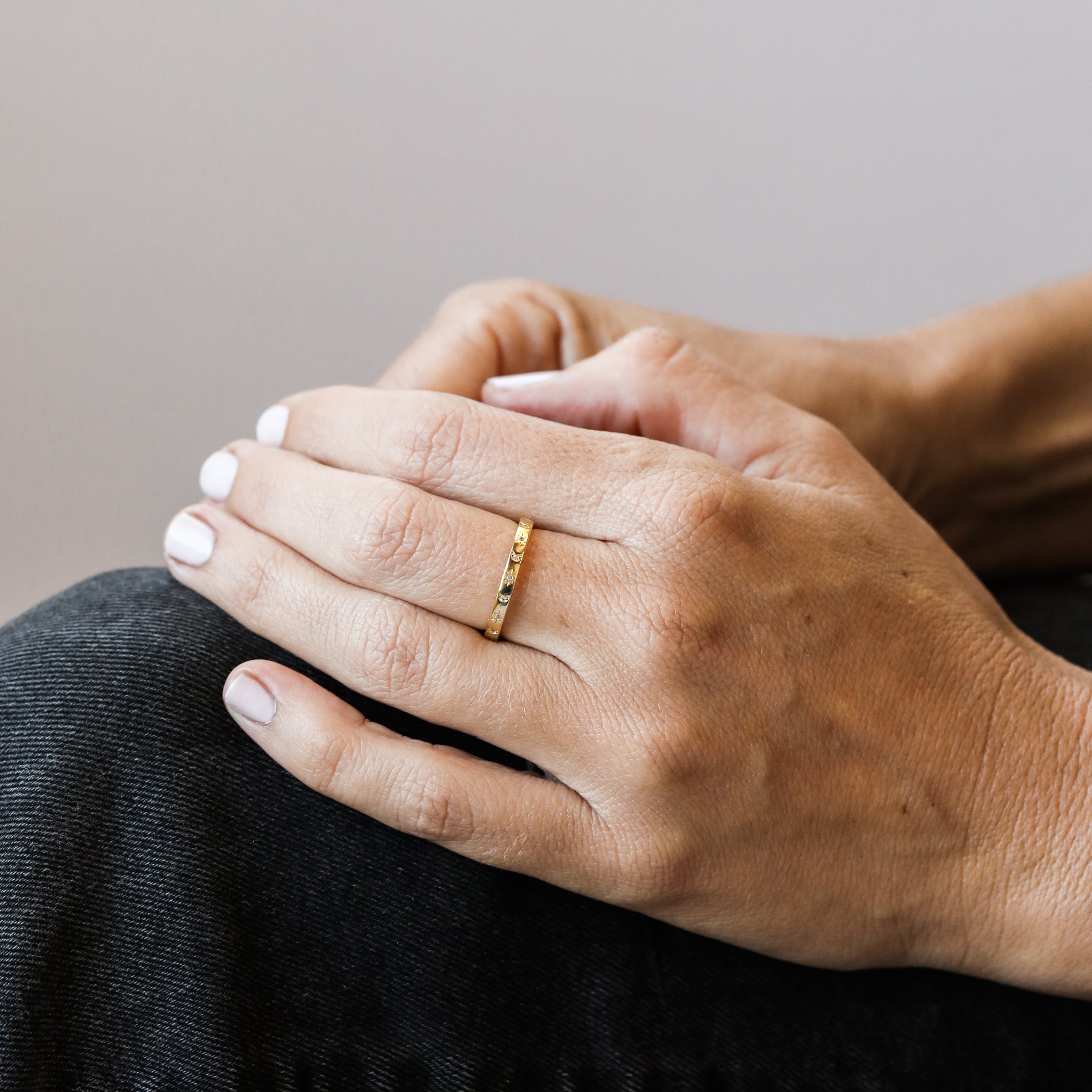 A close-up of a person’s hands on their knee, showing neatly manicured nails with white polish and the Celestial Ring—a simple gold stacking ring with delicate details—on one finger. The person is wearing dark pants.