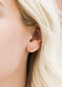 Close-up of a woman’s ear adorned with the Champagne Crawler: five small diamond studs in a line along her earlobe. Crafted with 18k gold plating and sterling silver posts, these earrings enhance her light skin and wavy blonde hair.