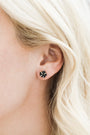 Close-up of a person with light blonde hair wearing the small, square-shaped Black Druzy Prong stud earring with 18k gold plating on their earlobe. The background and rest of the face are blurred.
