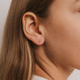 Close-up of a woman's ear with straight, light brown hair, wearing the Double Stud Stack—small gold studs featuring white baguette CZ. The neutral background is softly lit.