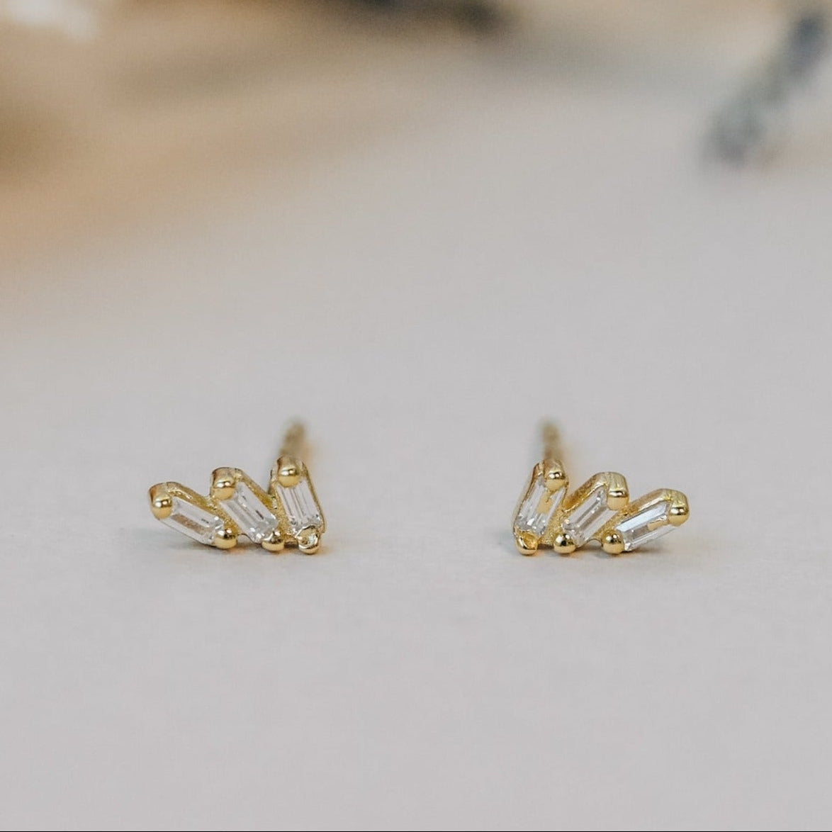 The Offset Trio - White CZ features delicate stud earrings with three baguette-cut white CZ stones in a fan shape, finished in 18k gold plating and presented on a neutral surface.