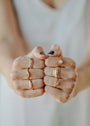 Close-up of two fists pressed together, featuring the Inset Baguette Ring and other gold rings on well-manicured fingers. The person wears a light sleeveless top, with one short nail painted black.