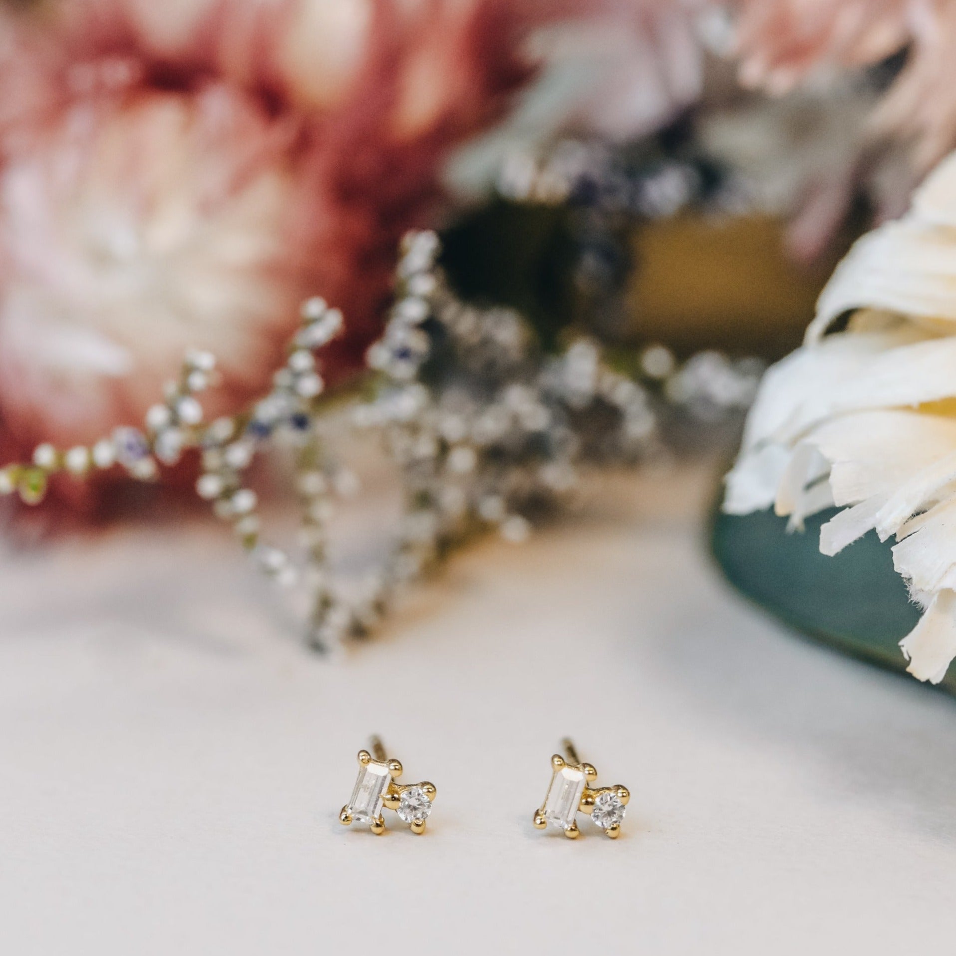 The Double Stud Stack earrings, featuring clear gemstones and 18k gold plating, are displayed on a light surface with softly blurred pink and white flowers in the background.