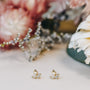 The Double Stud Stack earrings, featuring clear gemstones and 18k gold plating, are displayed on a light surface with softly blurred pink and white flowers in the background.