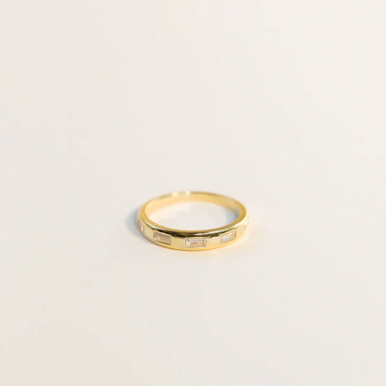 The Inset Baguette Ring is a thin gold band featuring small rectangular baguette-cut stones spaced evenly around it, shown on a simple light background.