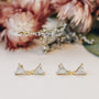 The Dual Triangle Drop features gold-plated earrings with two connected clear triangular stones and sterling silver posts, showcased on a light surface with softly blurred pink and beige flowers in the background.