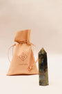 A tan suede JaxKelly pouch sits next to a polished Labradorite Crystal Point in blue and green tones, believed to create a protective aura, all displayed on a light beige background.