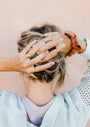 A woman with messy blonde hair, seen from behind, holds her head with both hands. She wears the Simple Stud Ring, a brown scrunchie, a light sweater, and a lavender top against a soft peach background.