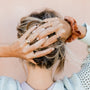 A woman with blonde hair in a messy bun holds her head, wearing the Lambda Ring and other gold rings, plus a brown scrunchie on her wrist. She wears a light sweater and stands against a pale pink background.